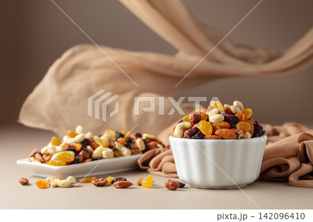 The mix of various nuts and raisins in a white bowl on a beige background. The mix of various nuts and raisins in a white bowl on a beige background. 142096410