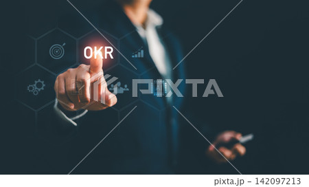Businessman touching glowing OKR text on futuristic screen with target graph icon. Concept for business goal management, objective key result, strategic planning, performance tracking, corporate Businessman touching glowing OKR text on futuristic screen with target graph icon. Concept for business goal management, objective key result, strategic planning, performance tracking, corporate 142097213
