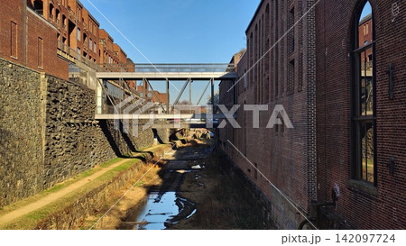 A cityscape featuring a metal bridge crossing historic brick buildings over a shallow canal in Georgetown. A clear sunny day highlights the strict geometry and texture of the walls. A cityscape featuring a metal bridge crossing historic brick buildings over a shallow canal in Georgetown. A clear sunny day highlights the strict geometry and texture of the walls. 142097724