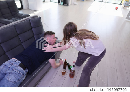 Woman helps intoxicated man off sofa in bright living room after drinking, concerned posture, scattered bottles on floor, daylight interior, domestic problem, support, recovery, safety, distress Woman helps intoxicated man off sofa in bright living room after drinking, concerned posture, scattered bottles on floor, daylight interior, domestic problem, support, recovery, safety, distress 142097793