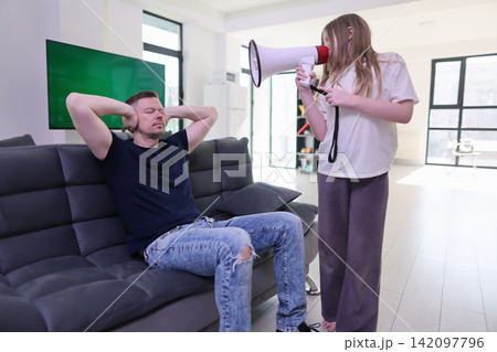 Young girl using megaphone beside relaxed man sitting on sofa in bright modern living room, expressing loud communication, attention, family tension, and domestic interaction at home Young girl using megaphone beside relaxed man sitting on sofa in bright modern living room, expressing loud communication, attention, family tension, and domestic interaction at home 142097796