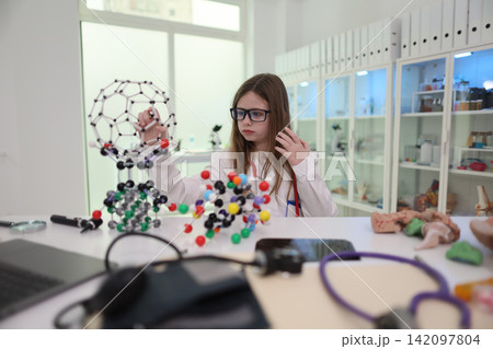 Young student in glasses studies colorful molecular model in bright science classroom with lab shelves, white coat, focused expression, educational STEM research environment, hands-on chemistry Young student in glasses studies colorful molecular model in bright science classroom with lab shelves, white coat, focused expression, educational STEM research environment, hands-on chemistry 142097804