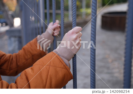 Child hands gripping metal fence bars outdoors in blurred playground setting, orange jacket sleeves and close up view suggesting curiosity, waiting, safety, confinement, and childhood activity Child hands gripping metal fence bars outdoors in blurred playground setting, orange jacket sleeves and close up view suggesting curiosity, waiting, safety, confinement, and childhood activity 142098254