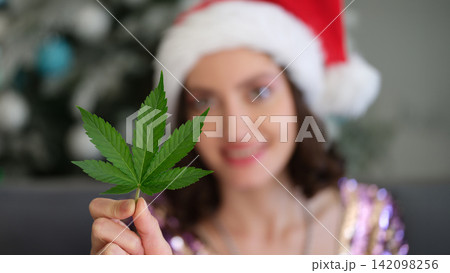 Smiling woman in Santa hat holding green cannabis leaf toward camera with festive holiday backdrop and soft blurred background, playful seasonal concept with unusual contrast and selective focus Smiling woman in Santa hat holding green cannabis leaf toward camera with festive holiday backdrop and soft blurred background, playful seasonal concept with unusual contrast and selective focus 142098256
