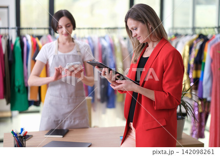 Two women reviewing fashion inventory in bright clothing showroom, one in red blazer checking clipboard while assistant in apron uses tablet beside colorful garment racks and workspace table Two women reviewing fashion inventory in bright clothing showroom, one in red blazer checking clipboard while assistant in apron uses tablet beside colorful garment racks and workspace table 142098261