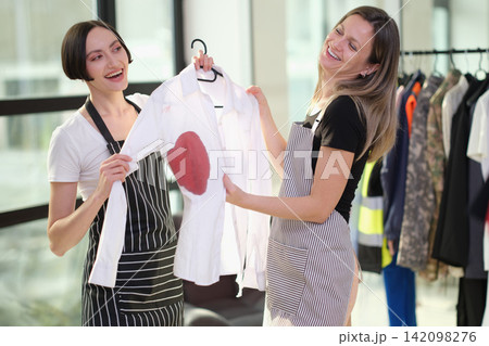 Two smiling women in aprons holding white shirt with red heart stain during laundry or garment care task beside clothing rack in bright studio with casual teamwork and creative repair concept 142098276
