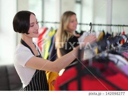 Smiling young woman browsing colorful clothing on hangers inside bright boutique while choosing garments for fashion shopping, with softly blurred shopper and racks in background Smiling young woman browsing colorful clothing on hangers inside bright boutique while choosing garments for fashion shopping, with softly blurred shopper and racks in background 142098277