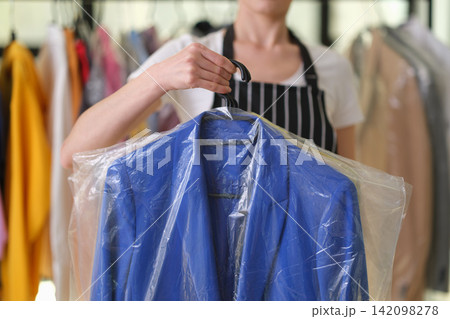 Person holding blue suit jacket inside clear garment cover inside clothing shop with blurred apparel racks behind, showing garment care, tailoring service, wardrobe management and retail fashion Person holding blue suit jacket inside clear garment cover inside clothing shop with blurred apparel racks behind, showing garment care, tailoring service, wardrobe management and retail fashion 142098278