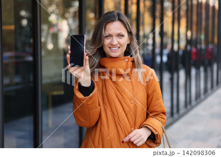 Smiling woman in orange quilted coat holding smartphone outdoors beside modern glass building with blurred city backdrop, presenting device screen toward viewer in urban lifestyle portrait Smiling woman in orange quilted coat holding smartphone outdoors beside modern glass building with blurred city backdrop, presenting device screen toward viewer in urban lifestyle portrait 142098306
