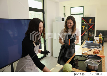 Two women preparing vegetables together in bright modern kitchen, smiling while cooking with cutting board, stovetop, sink, window light, organized shelves, fresh ingredients, and casual home 142098319