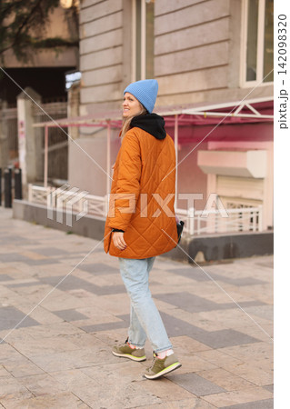 Smiling woman walking on city sidewalk wearing blue knit beanie, orange quilted coat, light jeans, and sporty sneakers, viewed from side against soft urban architecture Smiling woman walking on city sidewalk wearing blue knit beanie, orange quilted coat, light jeans, and sporty sneakers, viewed from side against soft urban architecture 142098320