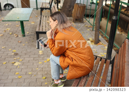 Woman in orange quilted coat sitting on park bench and checking mobile phone while resting among fallen autumn leaves in calm outdoor setting with reflective quiet mood Woman in orange quilted coat sitting on park bench and checking mobile phone while resting among fallen autumn leaves in calm outdoor setting with reflective quiet mood 142098323