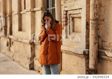 Young woman in orange quilted coat holding tissue to face beside weathered old wall on quiet street, showing seasonal discomfort and illness during cold weather outdoors Young woman in orange quilted coat holding tissue to face beside weathered old wall on quiet street, showing seasonal discomfort and illness during cold weather outdoors 142098324