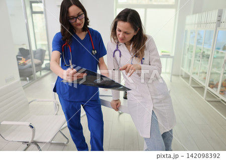 Two female doctors review chest radiograph together in bright modern hospital office, wearing scrubs and lab coats while discussing diagnosis beside chair and storage cabinets Two female doctors review chest radiograph together in bright modern hospital office, wearing scrubs and lab coats while discussing diagnosis beside chair and storage cabinets 142098392