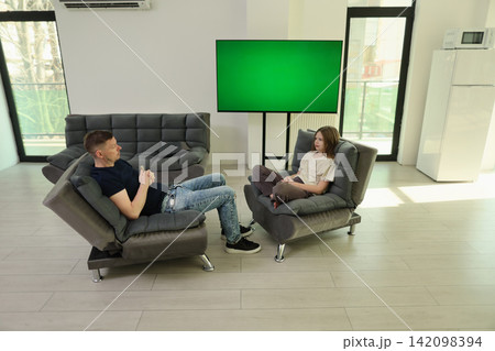 Man and young girl sitting across from each other in modern living room with green screen display, gray lounge chairs, bright daylight, casual indoor conversation setting Man and young girl sitting across from each other in modern living room with green screen display, gray lounge chairs, bright daylight, casual indoor conversation setting 142098394