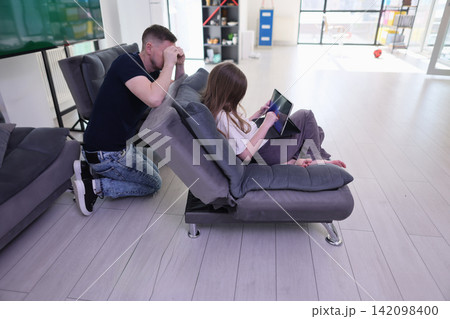 Father and young daughter spending quiet time on modern sofa while girl uses tablet and man kneels beside her in bright living room, showing concern, supervision and family attention at home Father and young daughter spending quiet time on modern sofa while girl uses tablet and man kneels beside her in bright living room, showing concern, supervision and family attention at home 142098400