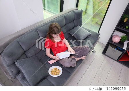 Young child reading book on gray sofa in bright modern home interior with smartphone and snack plate beside her, relaxed posture near large windows, shelving, cushions, and soft daylight 142099956