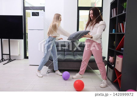 Two young women smiling while moving cushions in bright modern apartment, casual home scene with sofa, bookshelf, balloons, white walls, daylight, relaxed interior decorating and playful domestic Two young women smiling while moving cushions in bright modern apartment, casual home scene with sofa, bookshelf, balloons, white walls, daylight, relaxed interior decorating and playful domestic 142099972