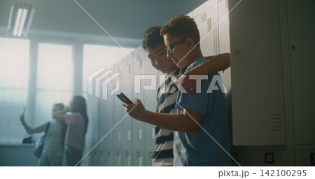 Friendly Teenage Classmates Relax by Lockers while One Shares Something Entertaining on Smartphone Friendly Teenage Classmates Relax by Lockers while One Shares Something Entertaining on Smartphone 142100295