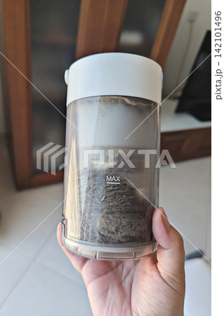Transparent vacuum dust bin filled with hair, lint and dirt, held by hand indoors before emptying and cleaning appliance Transparent vacuum dust bin filled with hair, lint and dirt, held by hand indoors before emptying and cleaning appliance 142101496