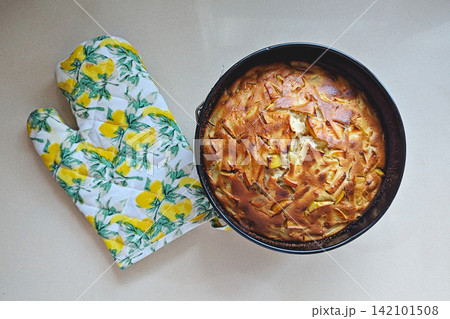Homemade apple sponge cake, traditional apple sharlotka, with baked apple pieces and golden crust in round pan beside oven mitt on light kitchen surface, top view 142101508