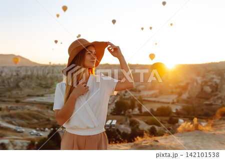 Attractive girl in hat stands on mountain with flying air balloons on background. Girl in sunrise. Attractive girl in hat stands on mountain with flying air balloons on background. Girl in sunrise. 142101538