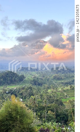 Mount Agung volcano peak rising above lush green tropical forests and rice paddies in Bali, Indonesia, with an airplane flying through the dramatic sunset clouds Mount Agung volcano peak rising above lush green tropical forests and rice paddies in Bali, Indonesia, with an airplane flying through the dramatic sunset clouds 142105675