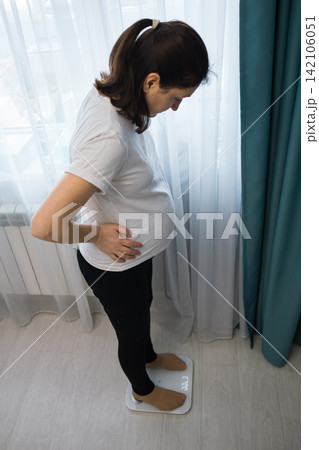 Pregnant woman standing on a digital scale in a bright room, checking her weight near sheer curtains and teal drapes, health and wellness scene at home Pregnant woman standing on a digital scale in a bright room, checking her weight near sheer curtains and teal drapes, health and wellness scene at home 142106051