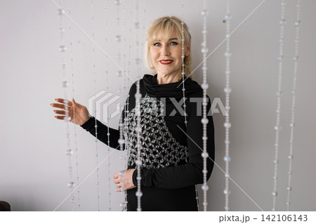 Elegant blonde woman smiling at the camera, wearing a black dress with a statement crystal necklace, reaching out to touch strands of a decorative beaded curtain Elegant blonde woman smiling at the camera, wearing a black dress with a statement crystal necklace, reaching out to touch strands of a decorative beaded curtain 142106143