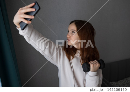 Young woman creating content for social media, capturing herself with a smartphone while holding a small dumbbell, showcasing her home workout and healthy lifestyle Young woman creating content for social media, capturing herself with a smartphone while holding a small dumbbell, showcasing her home workout and healthy lifestyle 142106150