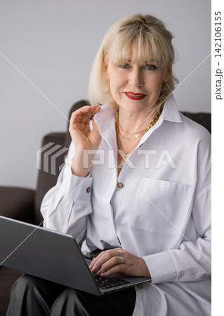 Elegant senior woman with blonde hair and red lipstick smiling confidently while typing on a laptop, representing remote work, online learning, and digital lifestyle 142106155