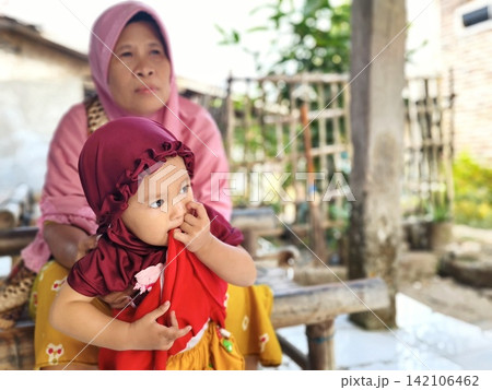 Indonesian grandmother and granddaughter wearing hijab sitting outside their village home. Indonesian grandmother and granddaughter wearing hijab sitting outside their village home. 142106462