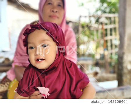 Indonesian grandmother and granddaughter wearing hijab sitting outside their village home. 142106464
