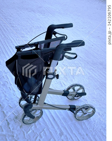 A foldable medical rollator for rehabilitation stands on a snow-covered road. A foldable medical rollator for rehabilitation stands on a snow-covered road. 142106795