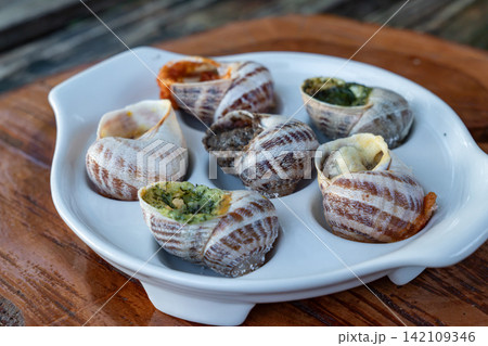 Baked stuffed mollusks are presented with green herb pesto and red spicy tomato sauces inside special white tray on timber background near metal fork outdoors. Baked stuffed mollusks are presented with green herb pesto and red spicy tomato sauces inside special white tray on timber background near metal fork outdoors. 142109346