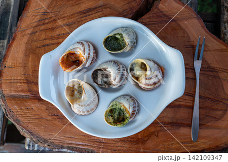 Baked stuffed mollusks are presented with green herb pesto and red spicy tomato sauces inside special white tray on timber background near metal fork outdoors. Baked stuffed mollusks are presented with green herb pesto and red spicy tomato sauces inside special white tray on timber background near metal fork outdoors. 142109347