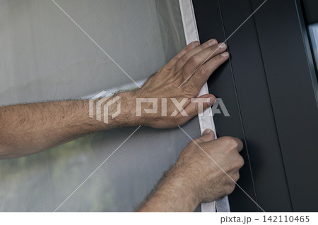 Hands securing a white mosquito screen into the window frame, illustrating precise handwork in home tasks and maintenance. Hands securing a white mosquito screen into the window frame, illustrating precise handwork in home tasks and maintenance. 142110465