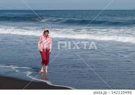 Woman stands barefoot at Pacific shoreline on black volcanic sand at Khalaktyrsky Beach, Kamchatka, Russia. Travel, nature tourism, coastal lifestyle, solitude, relaxation, summer vacation. Woman stands barefoot at Pacific shoreline on black volcanic sand at Khalaktyrsky Beach, Kamchatka, Russia. Travel, nature tourism, coastal lifestyle, solitude, relaxation, summer vacation. 142110499