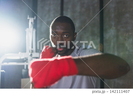 Workout in gym. African man fighter ready for fight punching with boxing wraps protective bandages to camera. Strong man training punches looking concentrated straight preparing for boxing sparring Workout in gym. African man fighter ready for fight punching with boxing wraps protective bandages to camera. Strong man training punches looking concentrated straight preparing for boxing sparring 142111407
