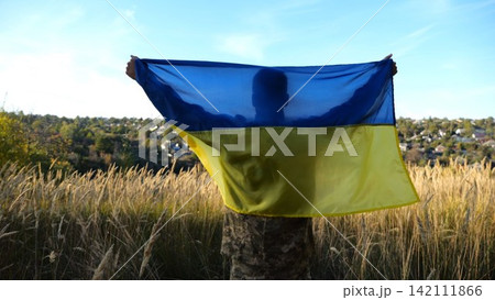 Military man in uniform stands with raised over head flag of Ukraine at countryside. Male ukrainian army soldier with lifted national banner in honor of victory against russian aggression. End of war 142111866