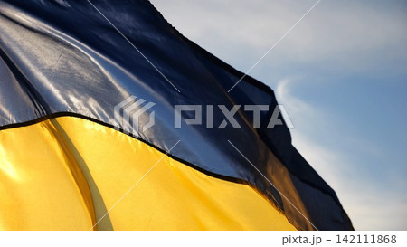 Blue-yellow symbol of Ukraine waving at wind against the background of sunlight. Ukrainian flag illuminated by sun fluttering at countryside. Concept of peace and support against russian aggression Blue-yellow symbol of Ukraine waving at wind against the background of sunlight. Ukrainian flag illuminated by sun fluttering at countryside. Concept of peace and support against russian aggression 142111868