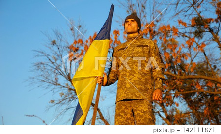 Ukrainian army man stands with national banner at countryside. Male soldier in military uniform with Ukraine flag as symbol of victory against russian aggression. Concept of invasion resistance Ukrainian army man stands with national banner at countryside. Male soldier in military uniform with Ukraine flag as symbol of victory against russian aggression. Concept of invasion resistance 142111871