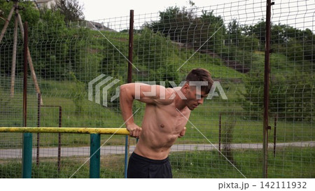 Young sportsman doing push ups on parallel bars at nature. Strong sporty man exercising at sports ground. Male shirtless athlete training hard outdoor. Concept of active sportive lifestyle. Slow mo Young sportsman doing push ups on parallel bars at nature. Strong sporty man exercising at sports ground. Male shirtless athlete training hard outdoor. Concept of active sportive lifestyle. Slow mo 142111932