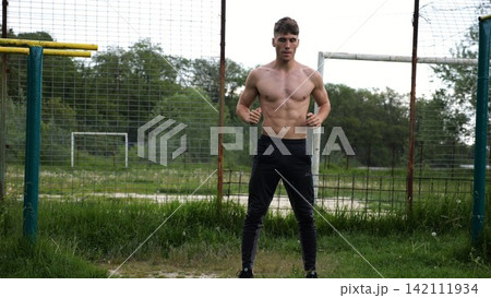 Male shirtless athlete doing squats at sports ground. Young sportsman working at summer nature. Strong sporty man exercising outdoor. Concept of active and sportive lifestyle. Slow motion Male shirtless athlete doing squats at sports ground. Young sportsman working at summer nature. Strong sporty man exercising outdoor. Concept of active and sportive lifestyle. Slow motion 142111934