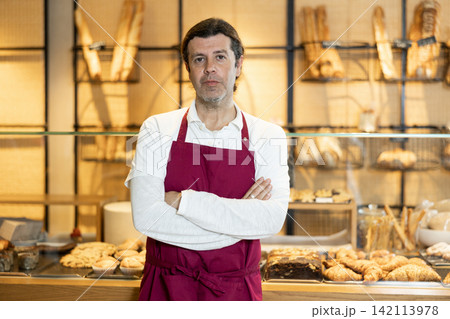 Positive adult bakery salesman in apron looking at the camera during working day in bakehouse. Cooking, culinary and bakery concept 142113978
