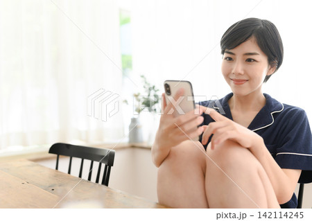 スマホを操作して微笑むパジャマ姿の女性 スマホを操作して微笑むパジャマ姿の女性 142114245