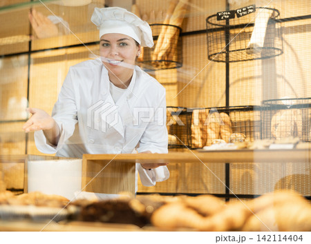Positive young female bakery worker in white toque and chef jacket selling pastry in bakeshop. Concept of small local business 142114404