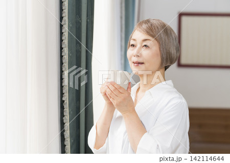 リビングの窓辺でコーヒーを飲むシニア女性 リビングの窓辺でコーヒーを飲むシニア女性 142114644