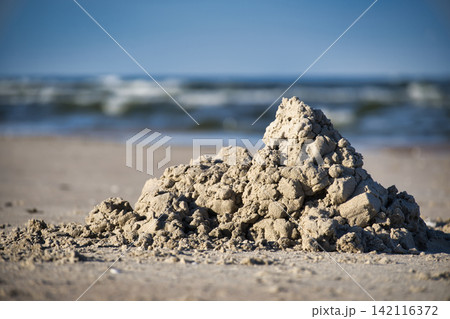Sandcastle Mound on Sandy Beach Shore with Blurred Ocean Waves and Blue Sky Background 142116372