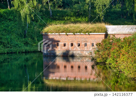 Historic Brick Fortification Wall with Green Grass Roof Reflected in Calm Water Surface 142116377
